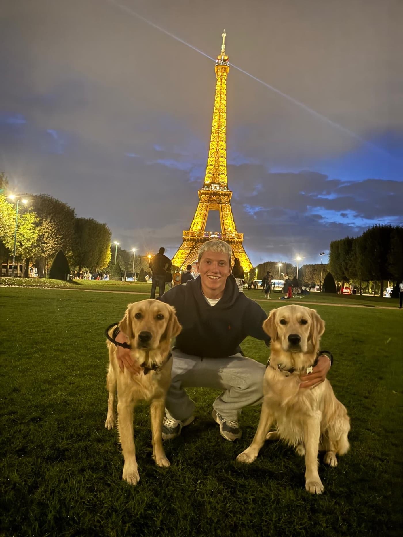 Seb crouched on the grass in front of the Eiffel Tower at night, a golden retriever either side of him